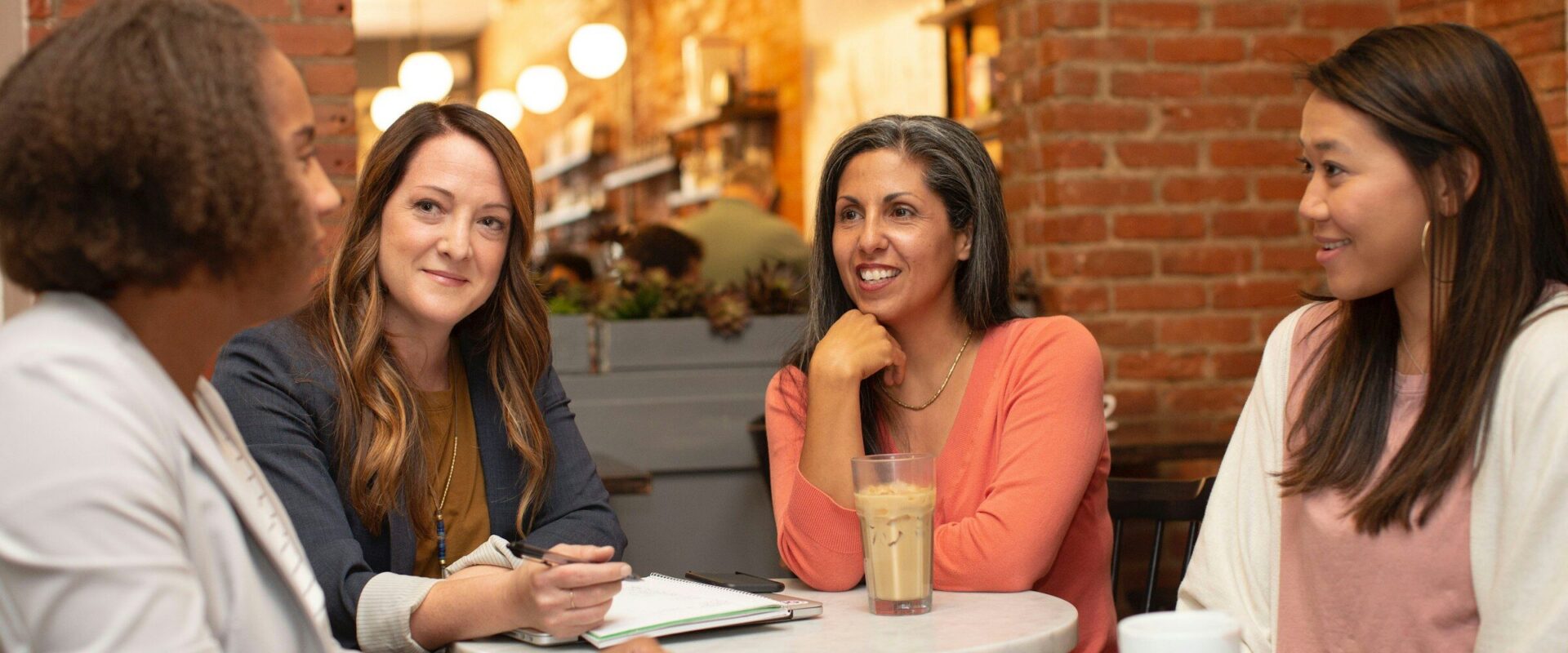 Four women in conversation around a small cafe table, one of them taking notes in a notebook.