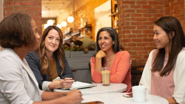 Four women in conversation around a small cafe table, one of them taking notes in a notebook.