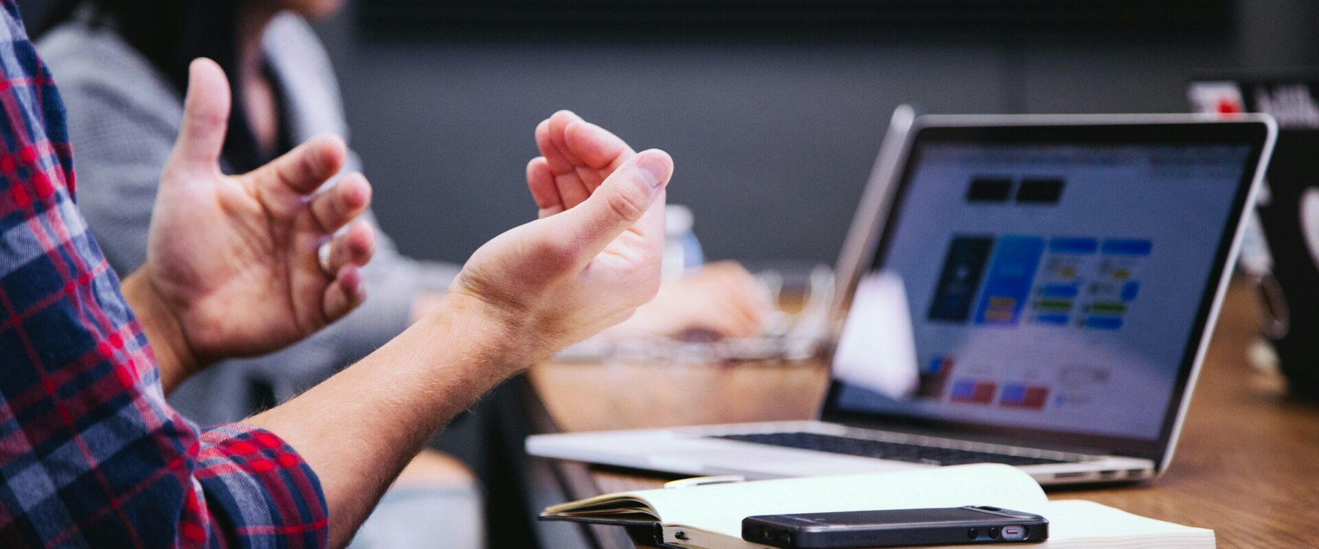 A person gesturing mid-explanation across a meeting table, with two open laptops showing dashboards.