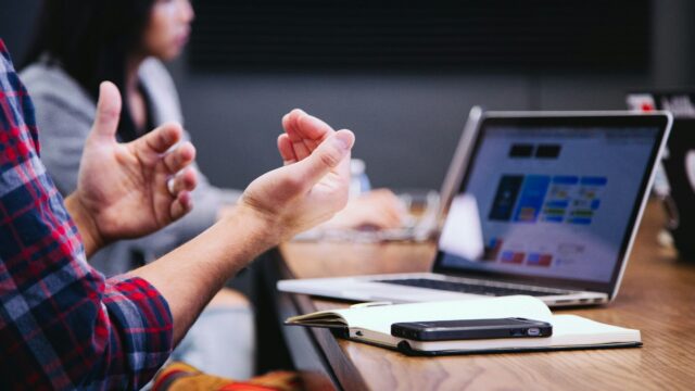 A person gesturing mid-explanation across a meeting table, with two open laptops showing dashboards.