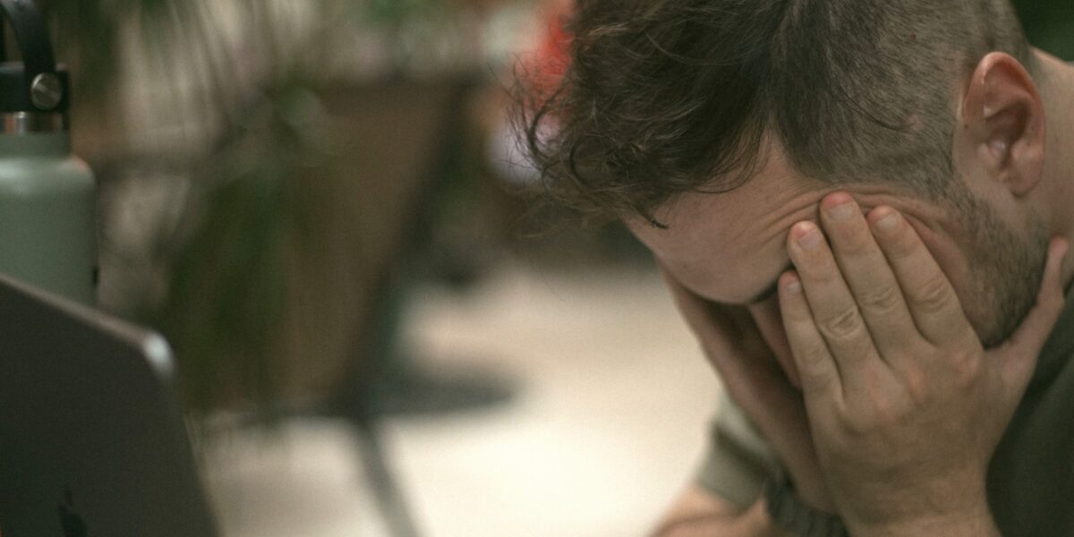 Frustrated man holding his head in his hands beside an open laptop.