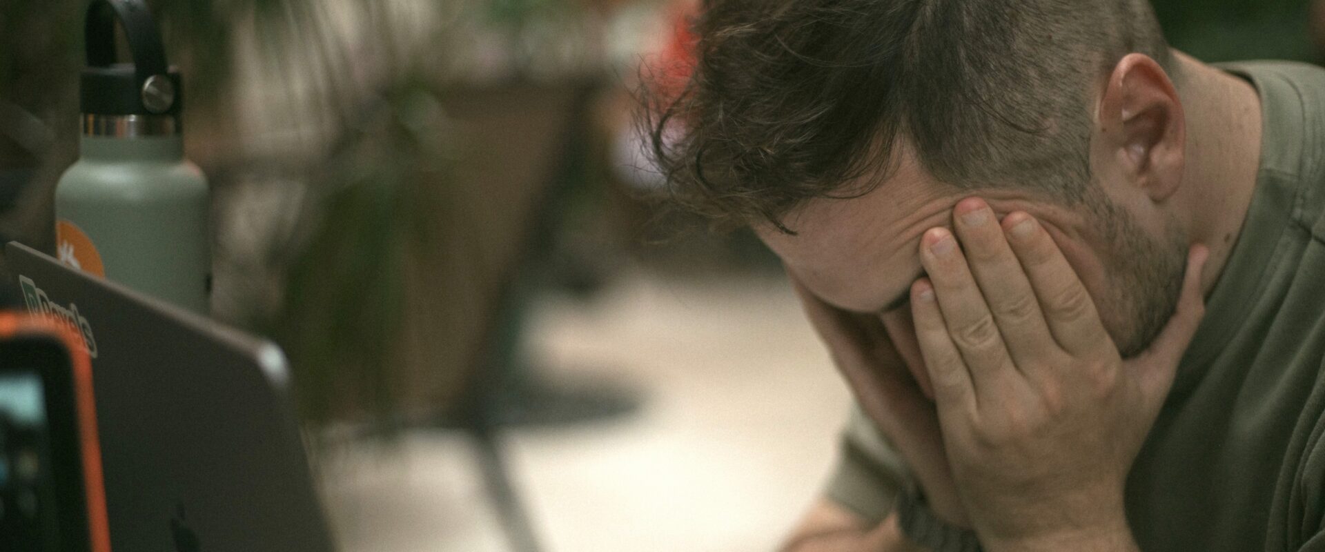 Frustrated man holding his head in his hands beside an open laptop.