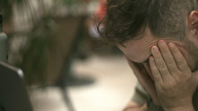Frustrated man holding his head in his hands beside an open laptop.